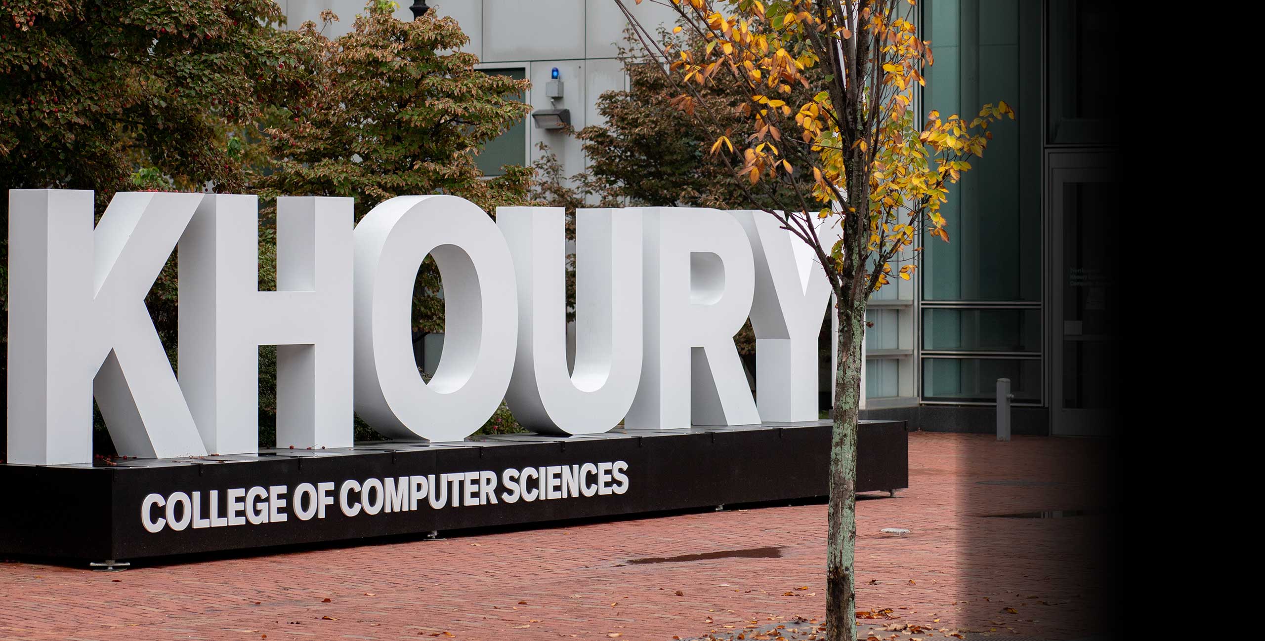 a photo showing the Khoury College of Computer Sciences sign outside of West Village H with fall foliage in the background