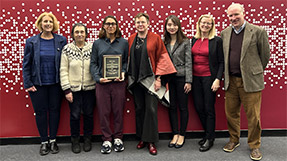 Kylie Bemis stands holding her award, surrounded by Khoury faculty and students