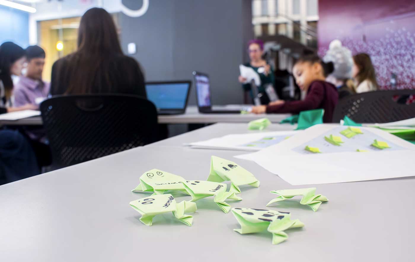 Six origami frogs made of bright green paper are scattered on a table while, in the background, Laney Strange speaks to the group of girls