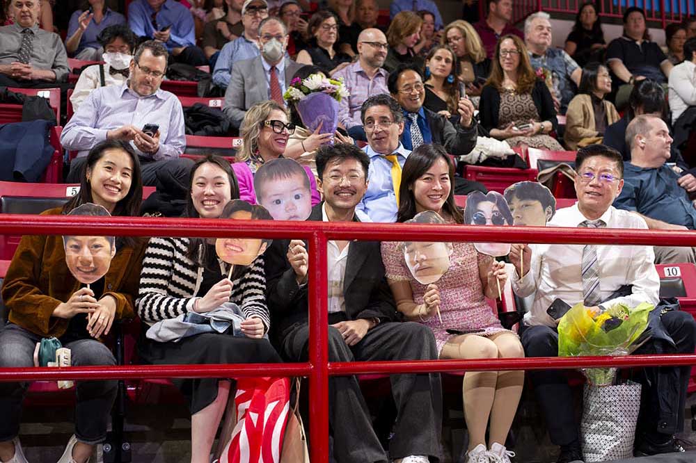 These guests found a fun way to show their Husky pride. A photo of a family of a graduate sitting in the audience at Matthews arena. The family members are holding up signs of a graduate's face attached to popsicle sticks.