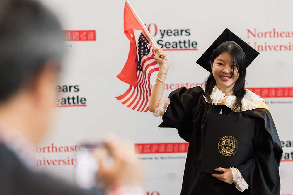 A graduate waves flags of the United States and China while holding her diploma at Commencement.