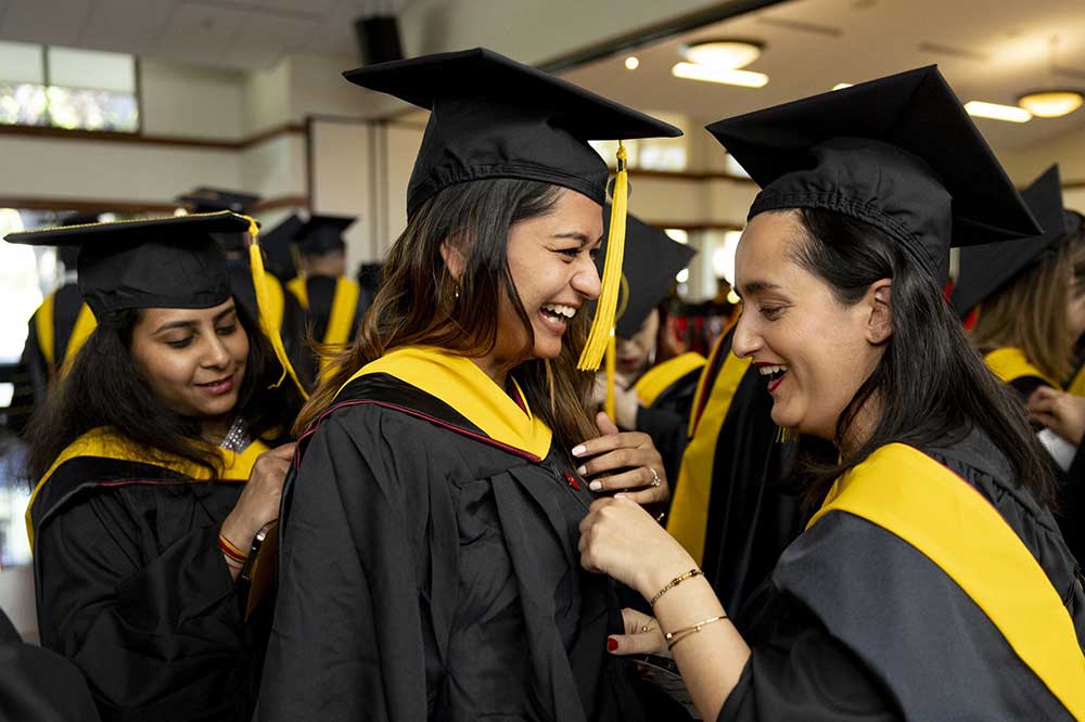 A graduate stands between two other graduates, who help her pin her robe and sash.
