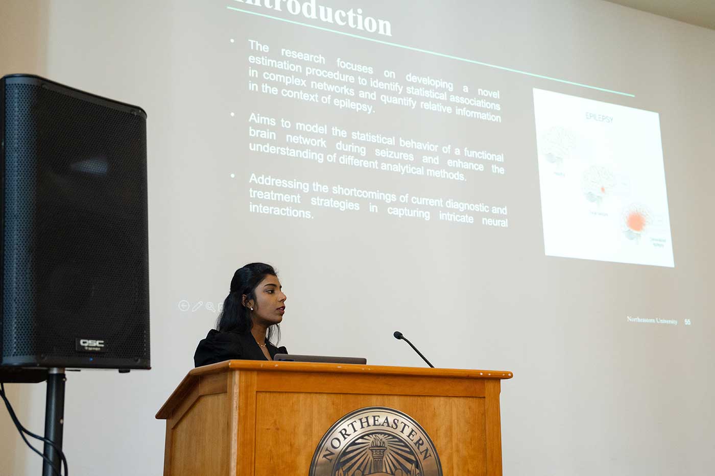 Divyadharshini Muruganandham presents a project while standing behind a podium, with the projection screen visible in the background