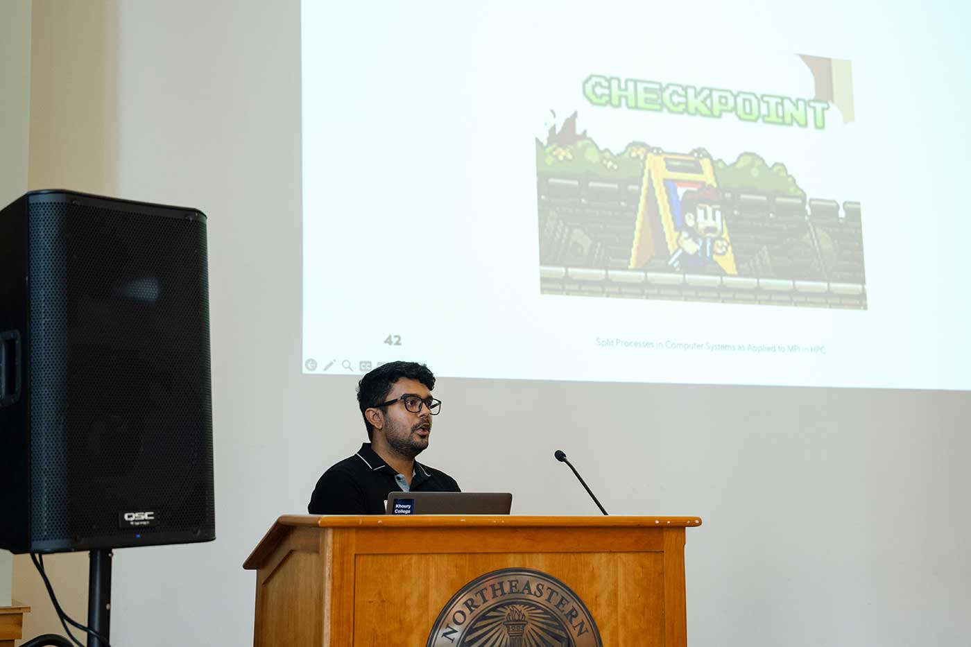 Kush Pandya presents a project while standing behind a podium, with the projection screen visible in the background