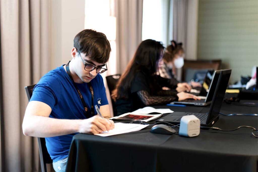 Three members of Nine members of Northeastern University’s Collegiate Cyber Defense Competition team sit at a large black table while working on a solution during a competition. The student on the left is writing on a piece of paper.