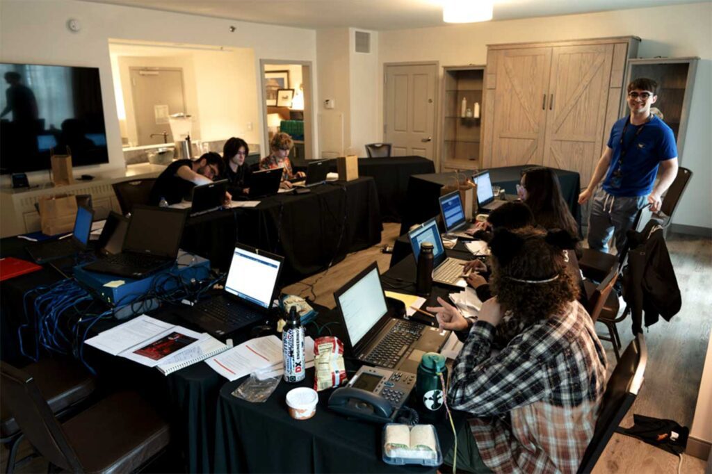 Six members of Nine members of Northeastern University’s Collegiate Cyber Defense Competition team sit at large tables and work on laptops during a competition.